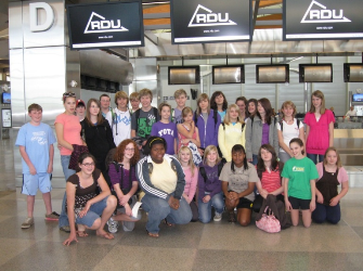 A group of students from a High School Exchange Program pose in an airport terminal, standing in front of RDU airport signs.